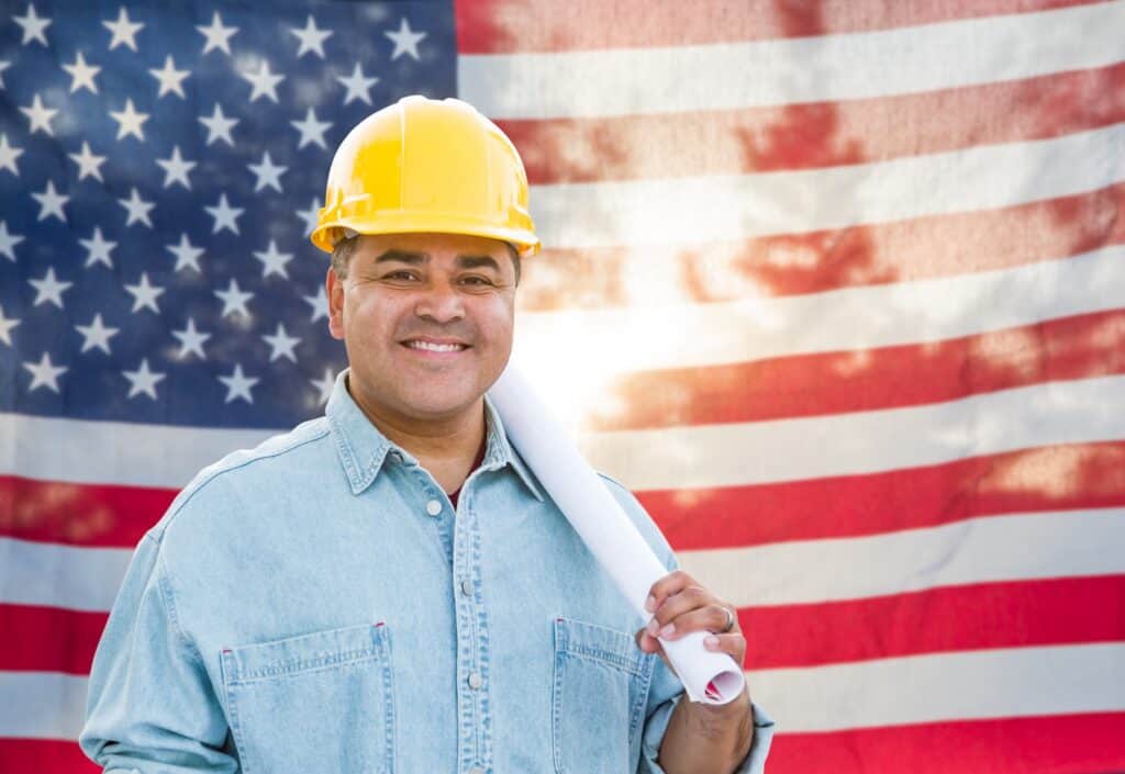 Hispanic male contractor with blueprint wearing hard hat in front of American Flag.