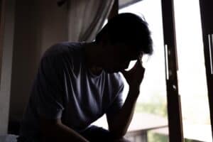 Anxious, overwhelmed adult man sitting in front of window and touching forehead