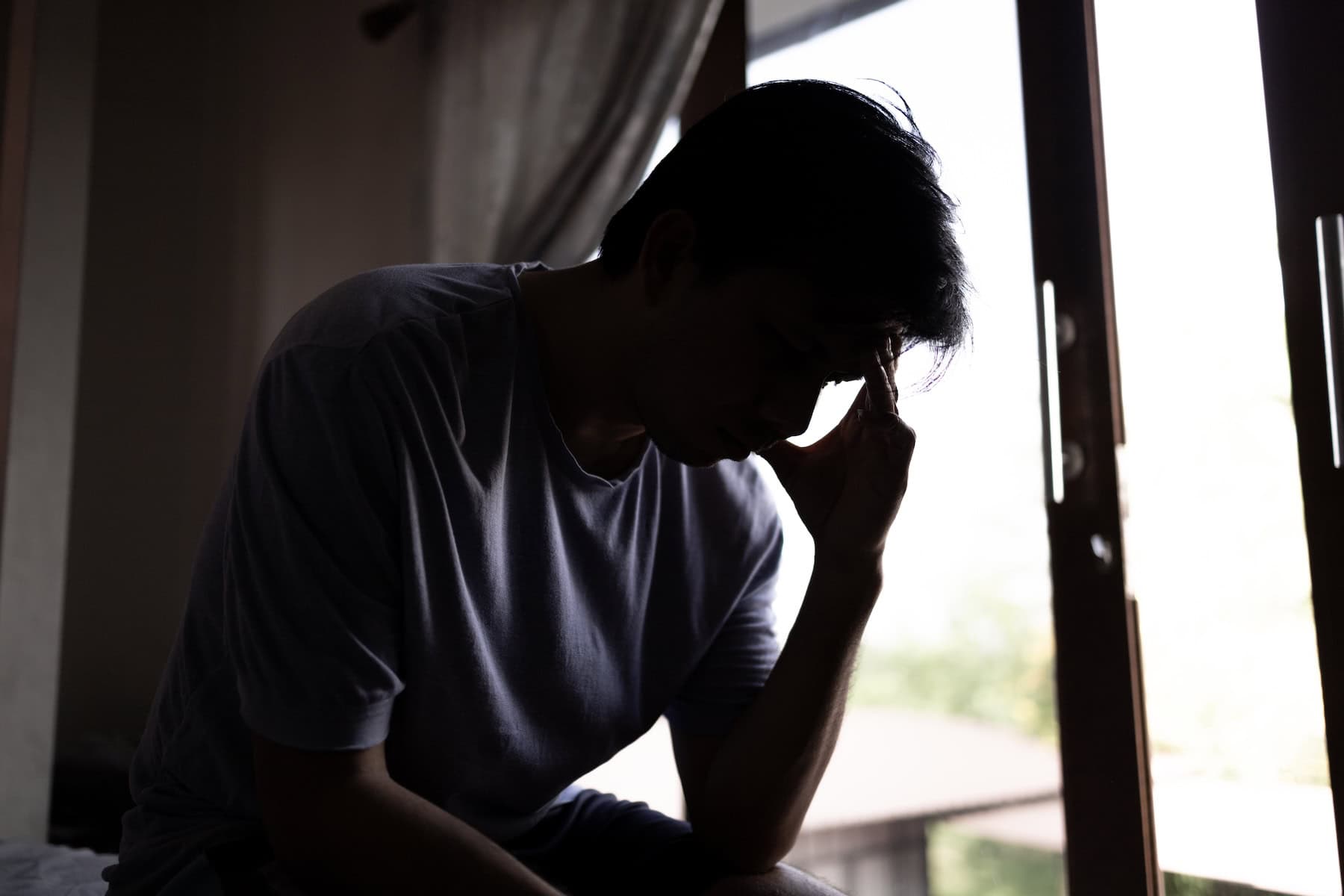 Hombre adulto ansioso y abrumado, sentado frente a una ventana y tocándose la frente.